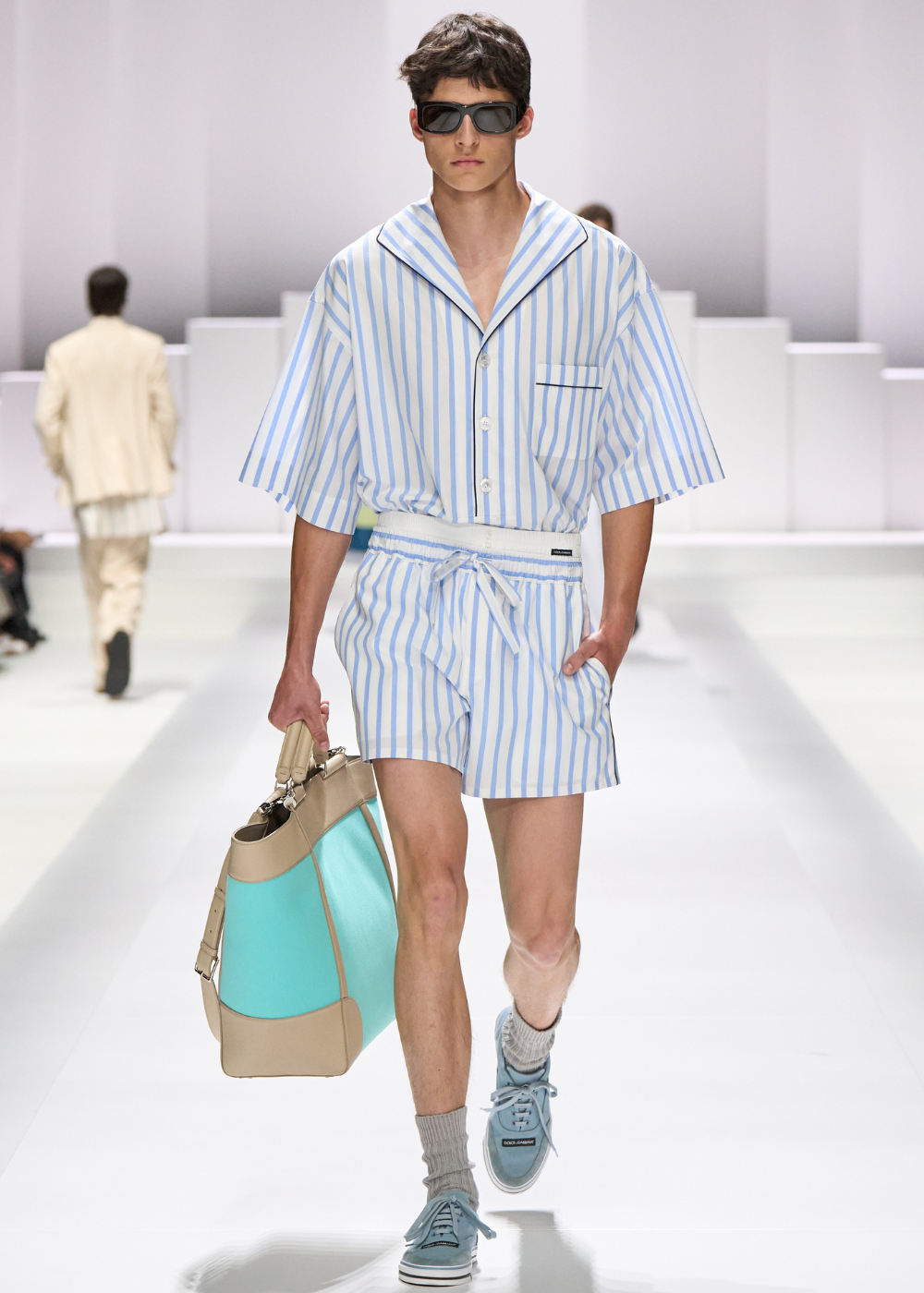 Modelo veste conjunto listrado azul estilo pijama, com bermuda e camisa de mangas curtas, carregando bolsa grande.