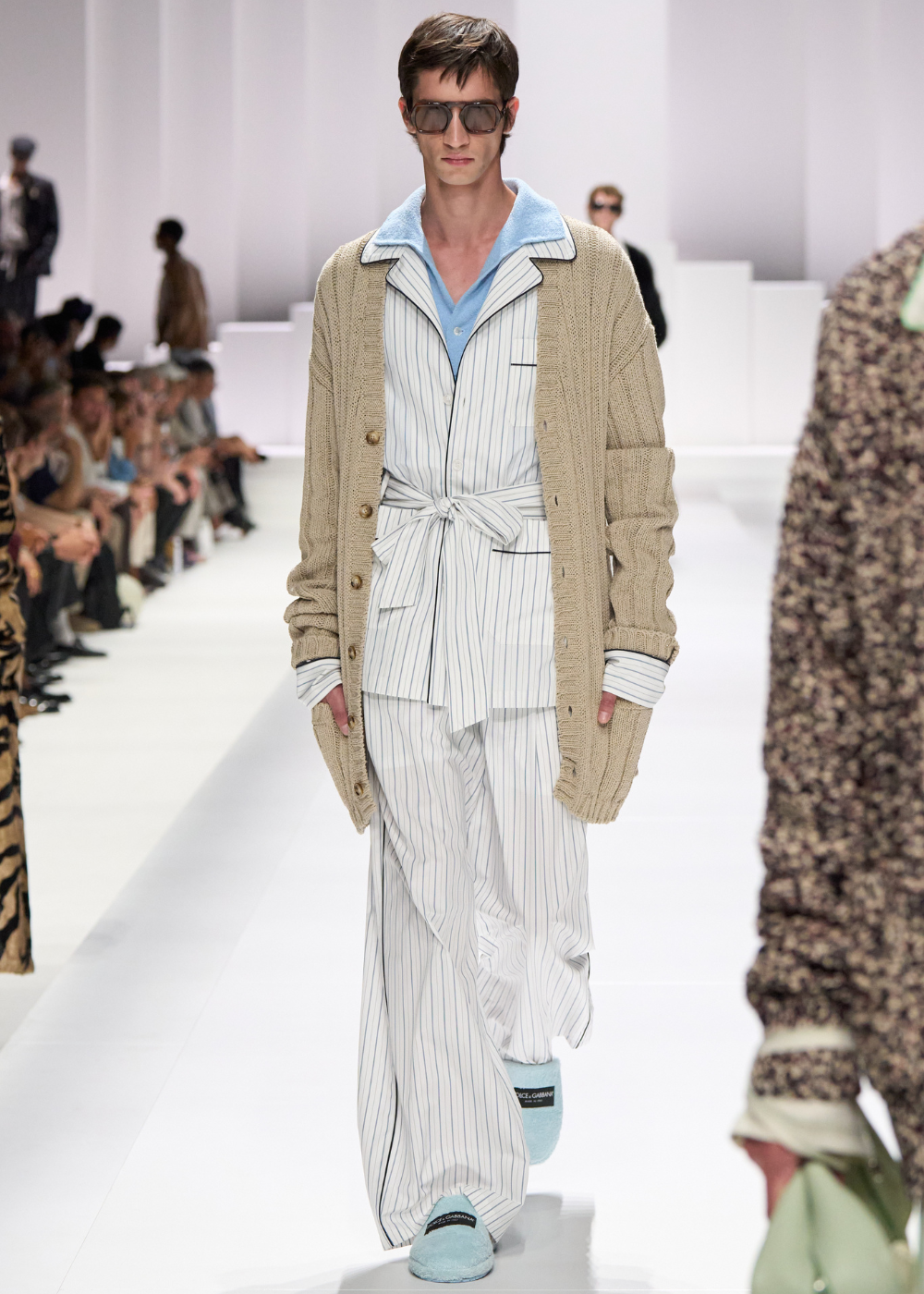 Modelo em desfile usando pijama listrado com roupão bege, óculos escuros e chinelos azuis.