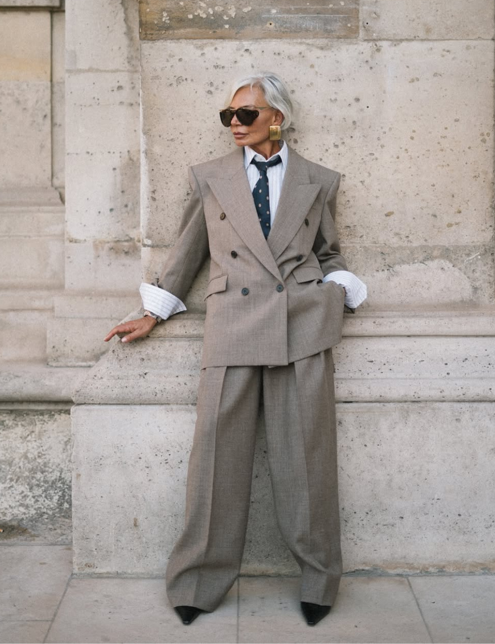 Senhora elegante com terno oversized cinza, exemplificando Power Dressing, junto a uma parede de pedra.