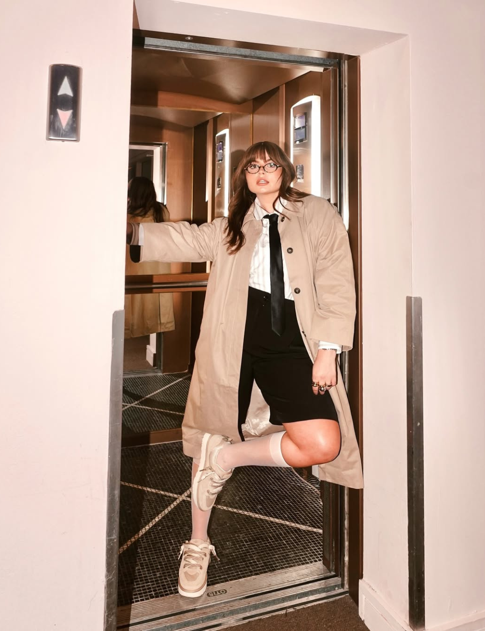 Pessoa com estilo Power Dressing em um elevador, usando casaco bege, camisa branca e tênis, transmite confiança e modernidade.