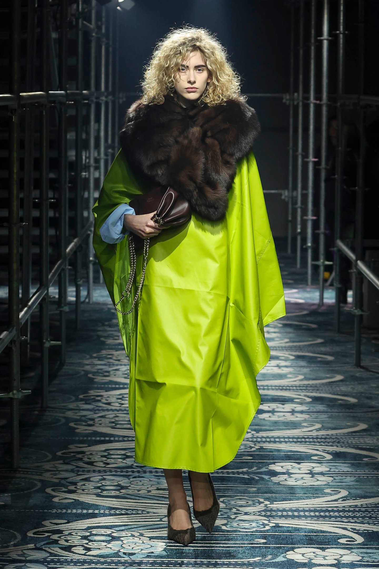 Modelo desfila em vestido verde tons neon com capa de pele, segurando bolsa marrom, em passarela industrial.