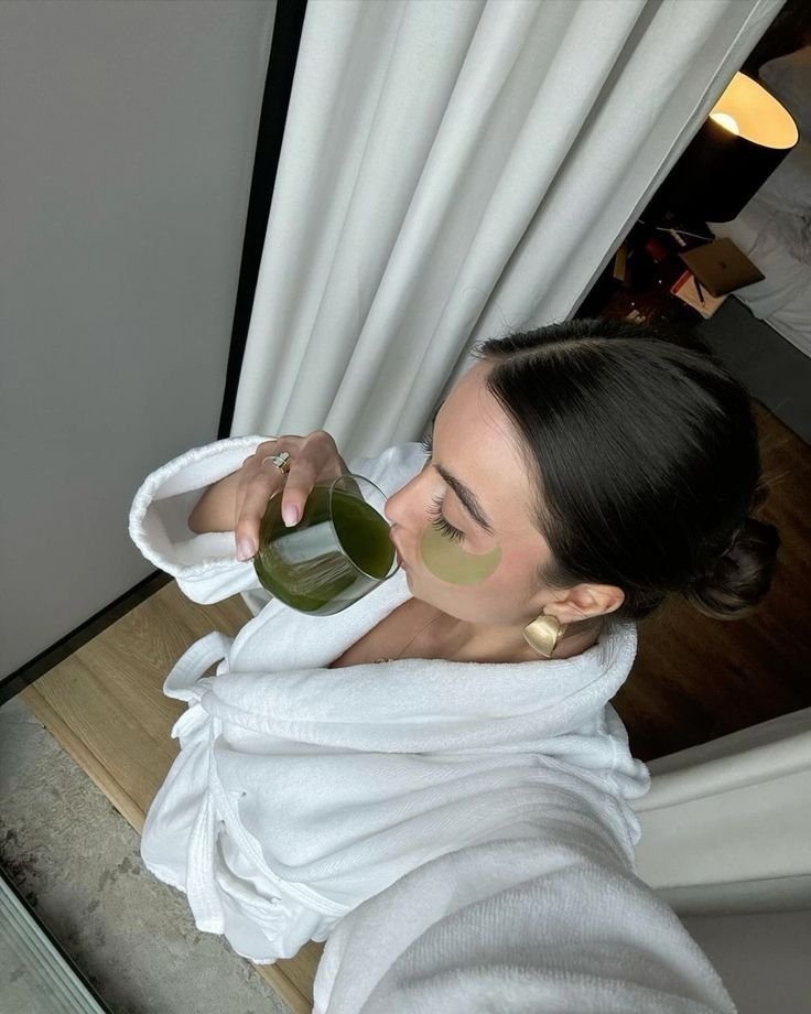 Mulher em roupão branco com máscara facial e suco verde, simbolizando procedimentos estéticos em alta.
