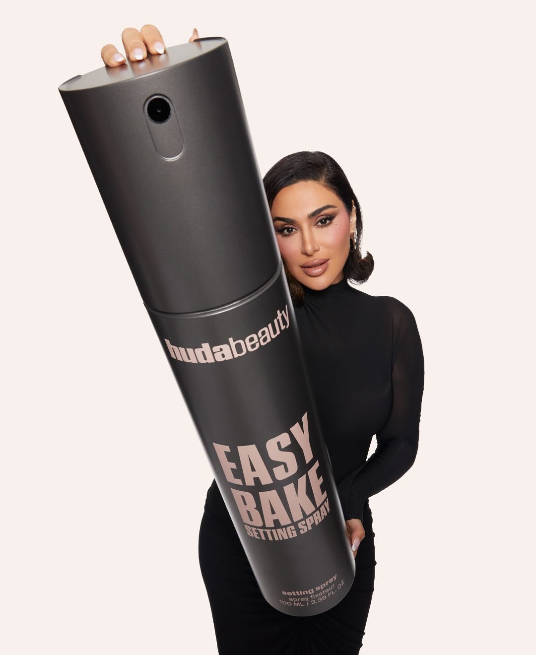 Mulher segura spray gigante da Huda Beauty, vestindo preto. A palavra-chave