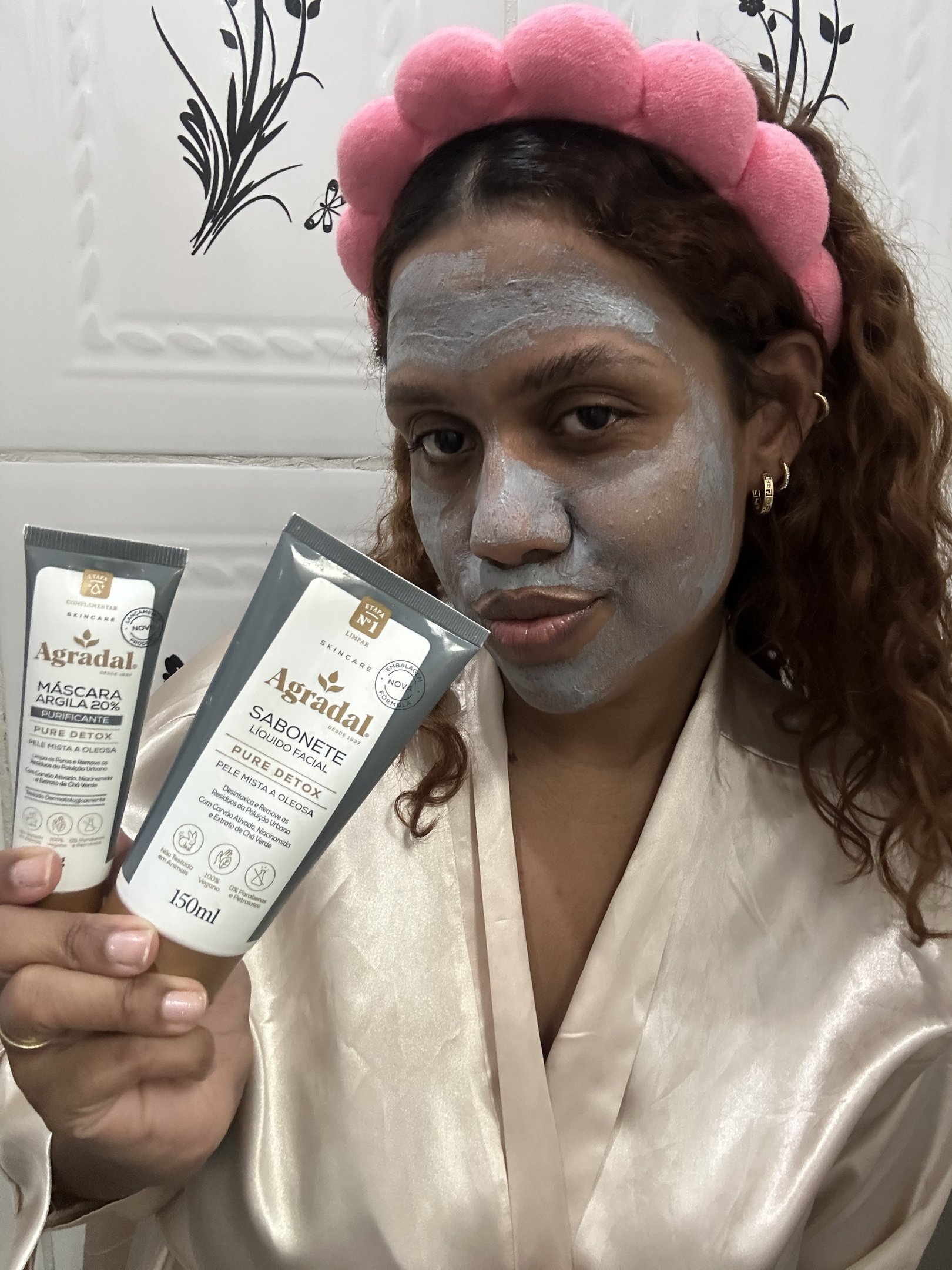 Pessoa com máscara argila no rosto segura produtos de cuidado facial, usando uma tiara rosa.