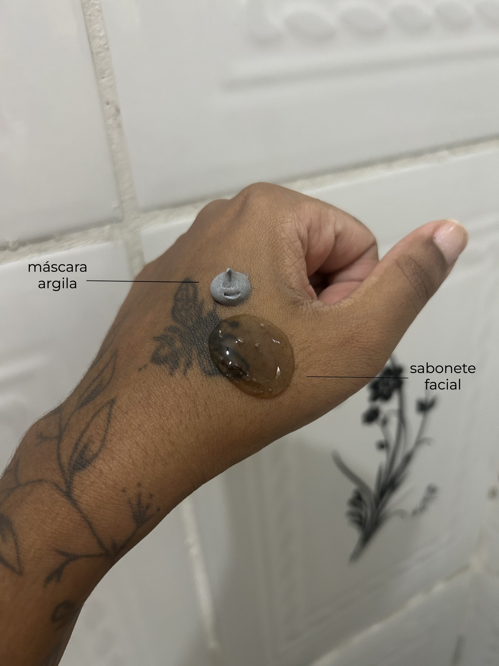 Mão com tatuagem, contendo máscara argila cinza e sabonete facial, sobre fundo de azulejo.