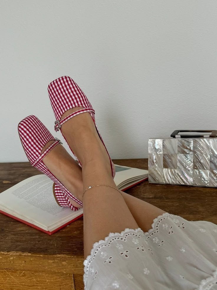 Sapatos xadrez vermelho e branco com laço, salto bloco, pernas cruzadas sobre livro aberto e bolsa decorativa.