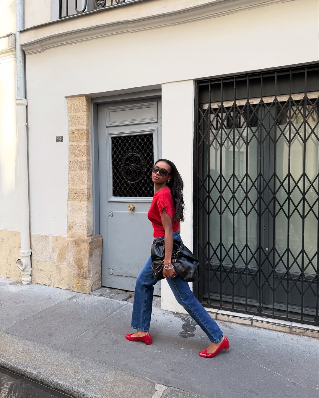 Mulher de blusa vermelha, calça jeans e scarpin salto bloco caminha em rua ao lado de portão cinza.