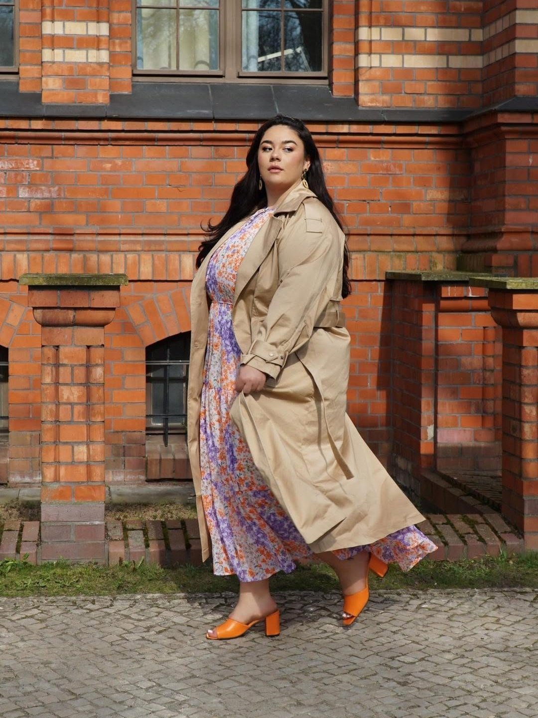 Pessoa caminha usando vestido floral com casaco bege e sandálias laranja de salto bloco, em frente a um prédio de tijolos.