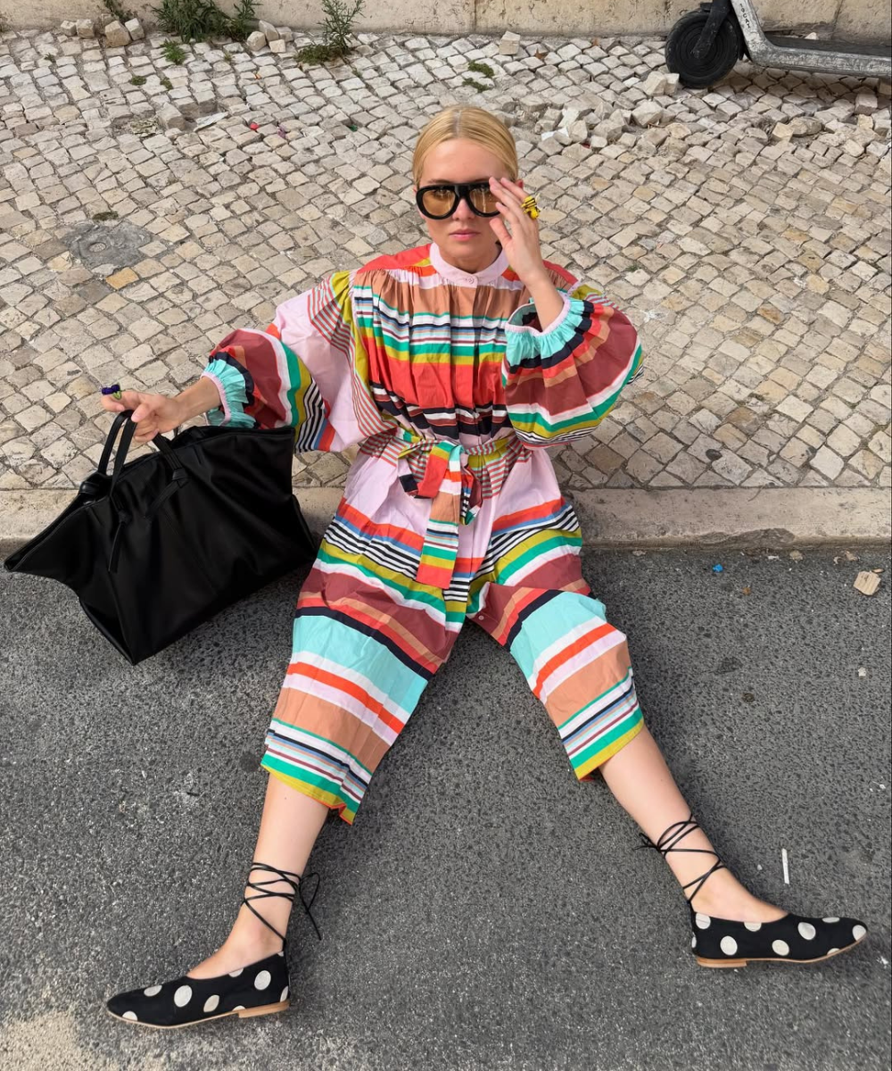 Pessoa sentada no chão, vestindo roupa colorida e sapatilha de amarração preta com bolinhas.