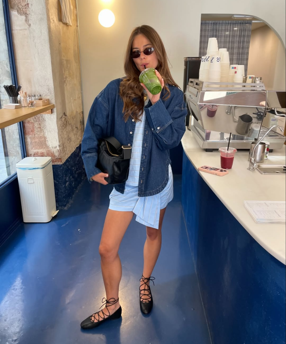 Mulher usando sapatilha de amarração preta e jaqueta jeans, bebe suco verde em uma cafeteria moderna.