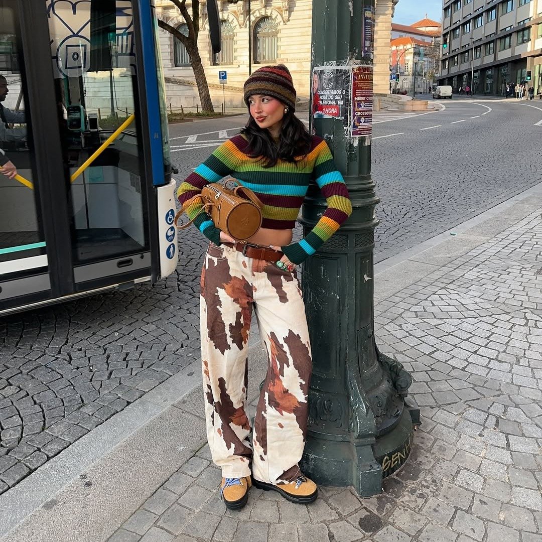 Pessoa com blusa listrada e calça estampada segura bolsa, usa sapato marrom em frente a um poste.