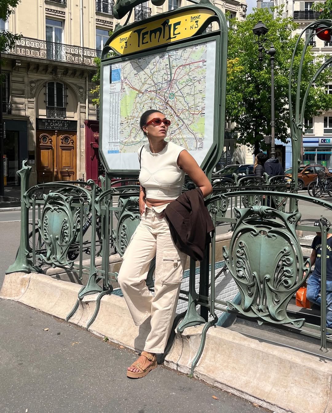 Pessoa de óculos está de pé com roupas casuais perto de uma entrada de metrô em Paris, usando sapatos de plataforma.
