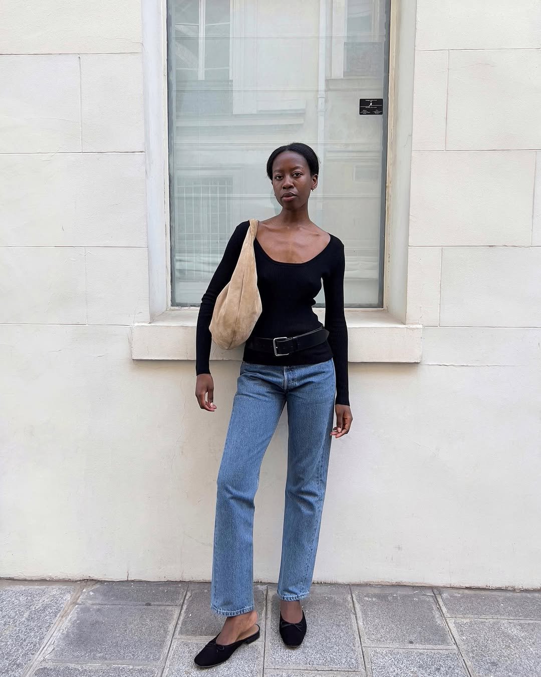 Pessoa em pé, com blusa preta e jeans, usando mule feminino preto e uma bolsa bege, em frente a uma parede clara.