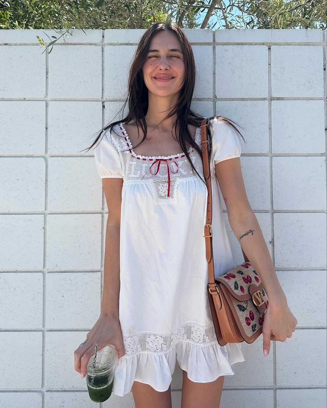 Jovem com vestido branco sorrindo ao ar livre, segurando bebida verde e bolsa estampada.