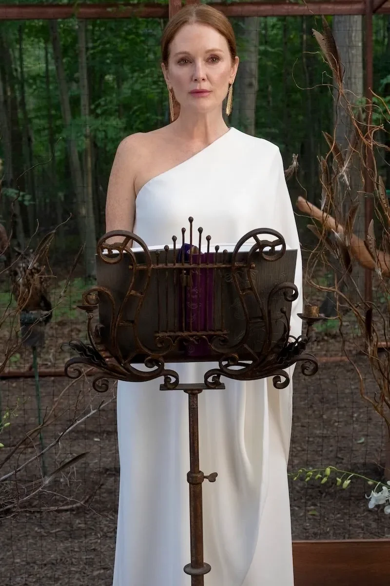 Uma mulher de vestido branco está em frente a um suporte ornamentado, cercada por árvores. Sereias