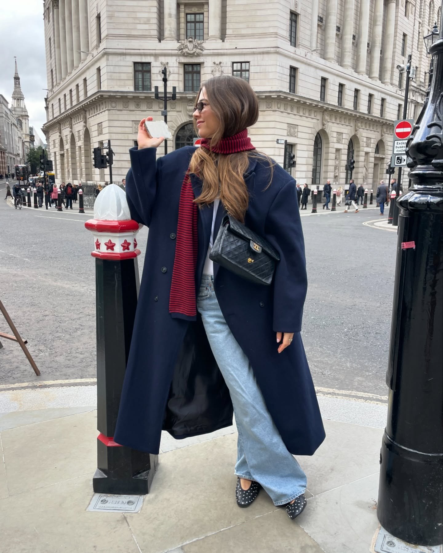 Mulher de sobretudo feminino azul, cachecol vermelho, calça jeans e bolsa preta, posa em rua movimentada.