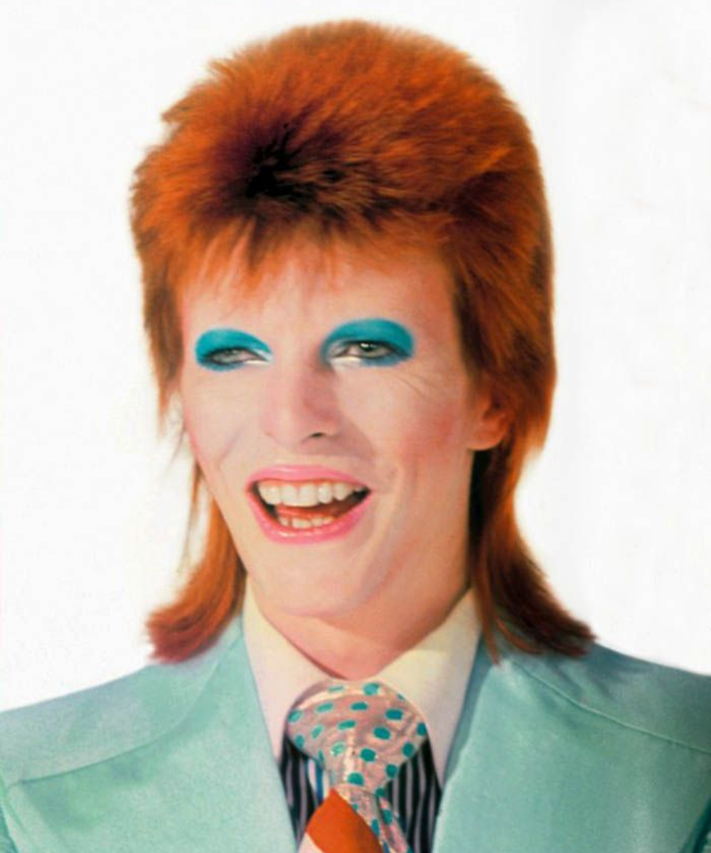 Pessoa sorridente com cabelo alaranjado e maquiagem marcante. Olhos com sombra azul. Usa terno azul claro e gravata estampada.