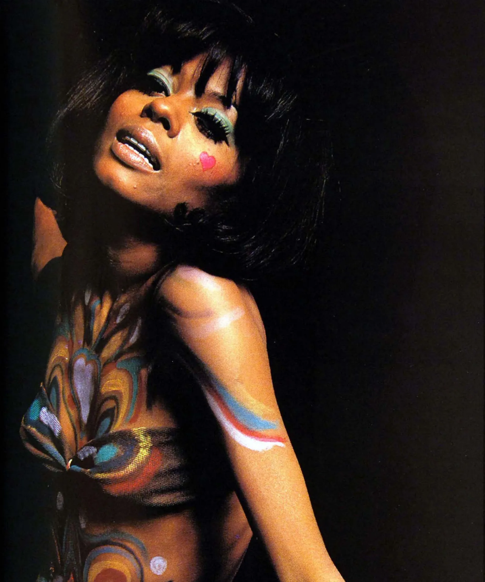 Rosto maquiado com sombra azul e coração pintado na bochecha, corpo coberto de pintura artística colorida.