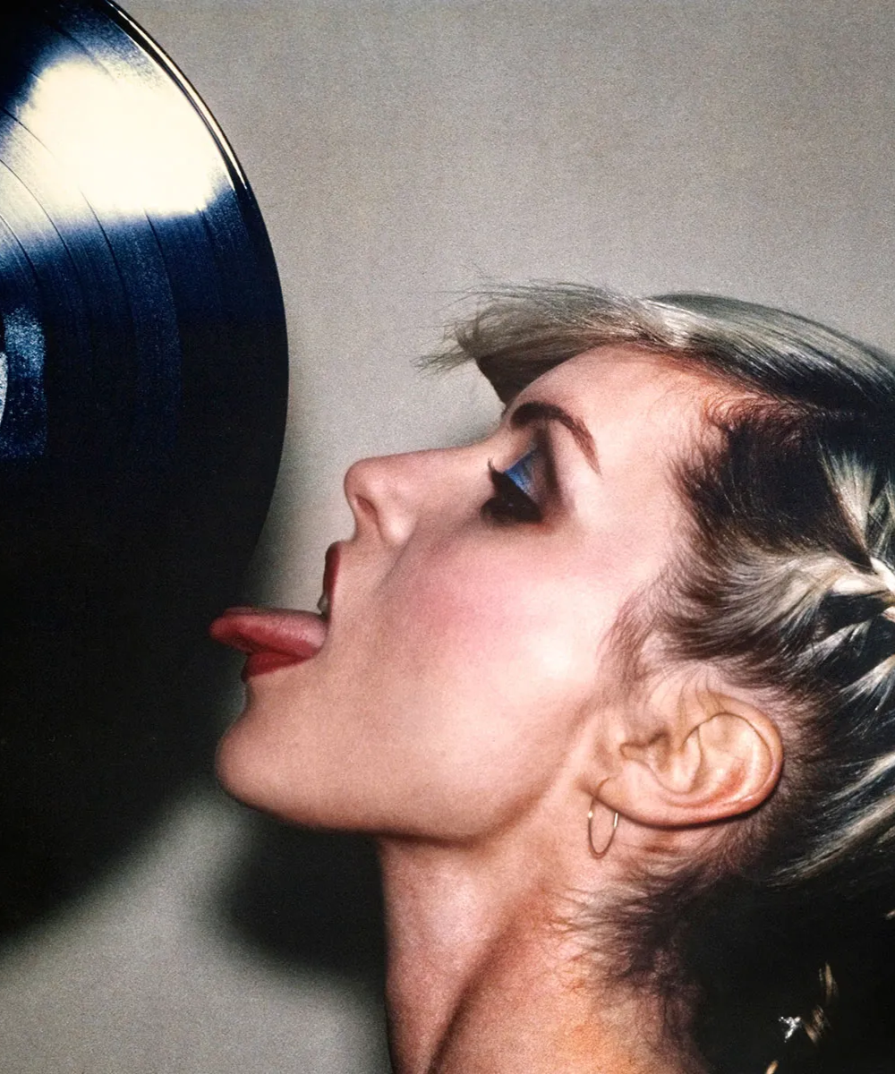 Mulher com maquiagem sombra azul e batom vermelho lambe um disco de vinil refletindo luz.