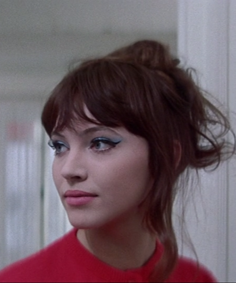 Jovem com sombra azul nos olhos e cabelo castanho preso, veste suéter vermelho. Ela olha para o lado.