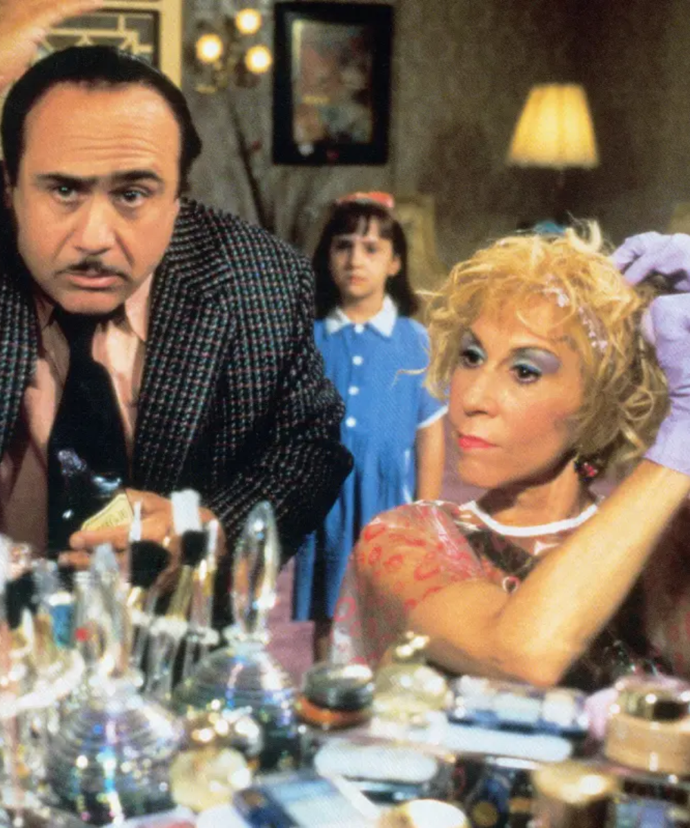 Homem e mulher arrumando-se em frente ao espelho, com sombra azul nas pálpebras, enquanto criança observa ao fundo.