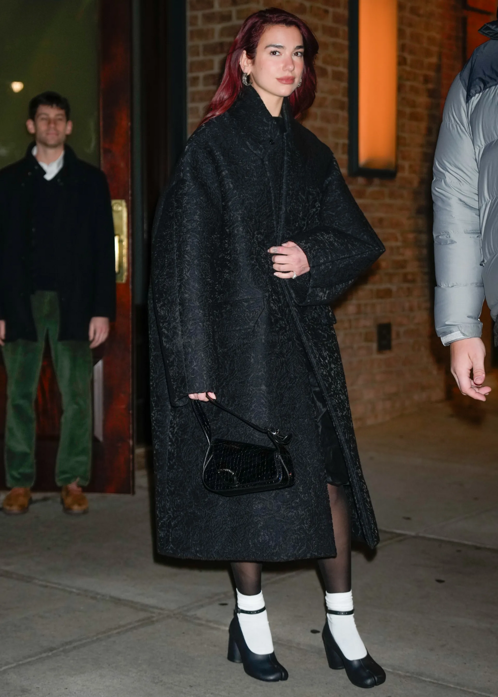 Pessoa andando com casaco longo preto e sapatos de salto usados por celebridades, em rua à noite.