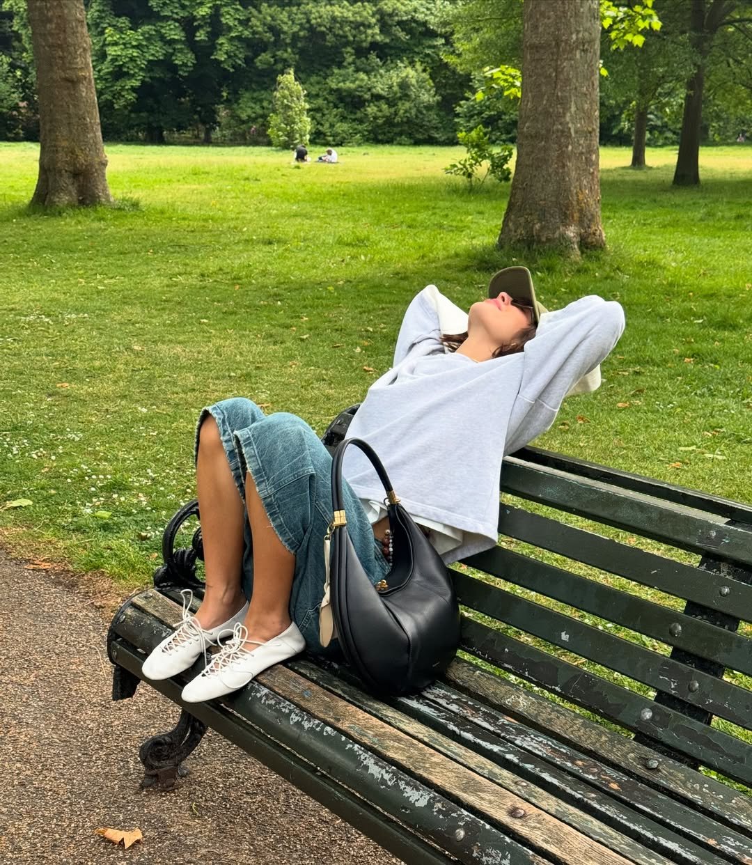 Mulher relaxa em banco de parque, usando sapatilhas brancas modernas e saia jeans, em meio a árvores e grama.
