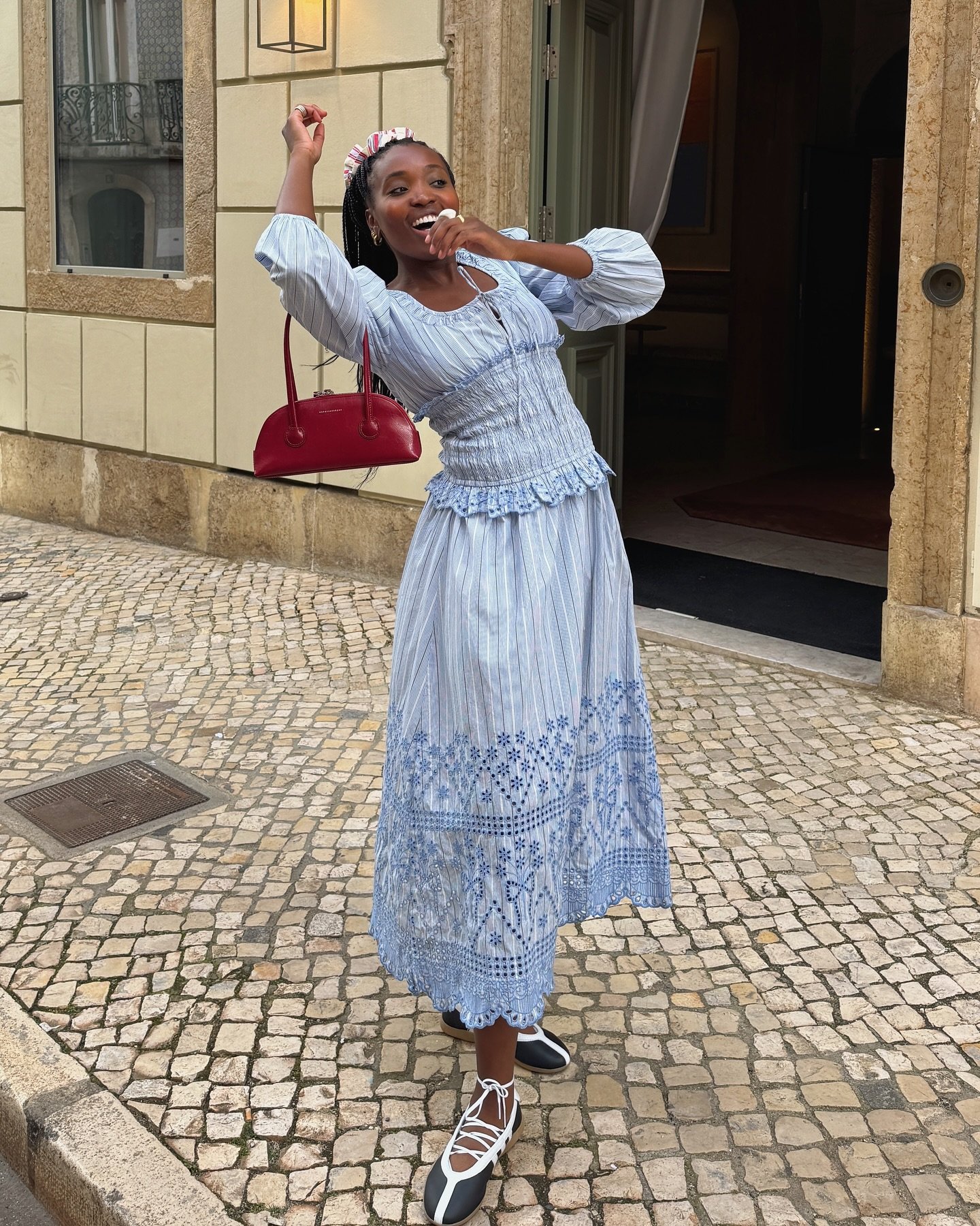 Mulher sorrindo, com vestido azul e sapatilhas estilosas, posa em rua de paralelepípedos.