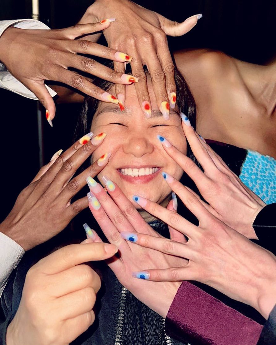 Diversas mãos com unhas decoradas em estilo coreano com cores vibrantes cercam um rosto sorridente.