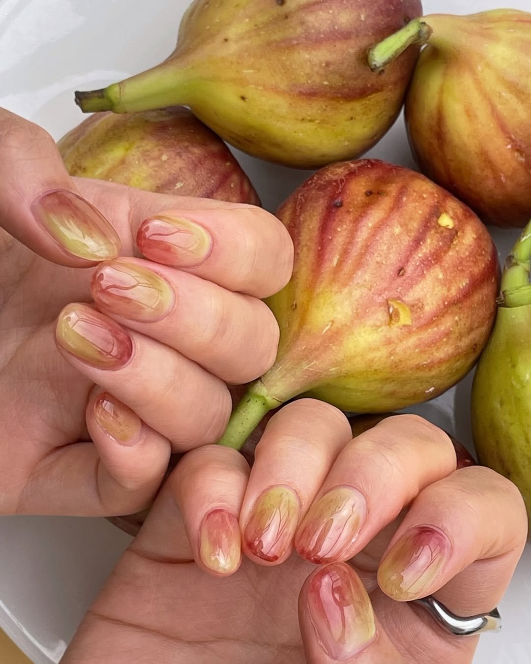 Unhas decoradas em tons de verde e vermelho, combinando com figos maduros ao fundo.