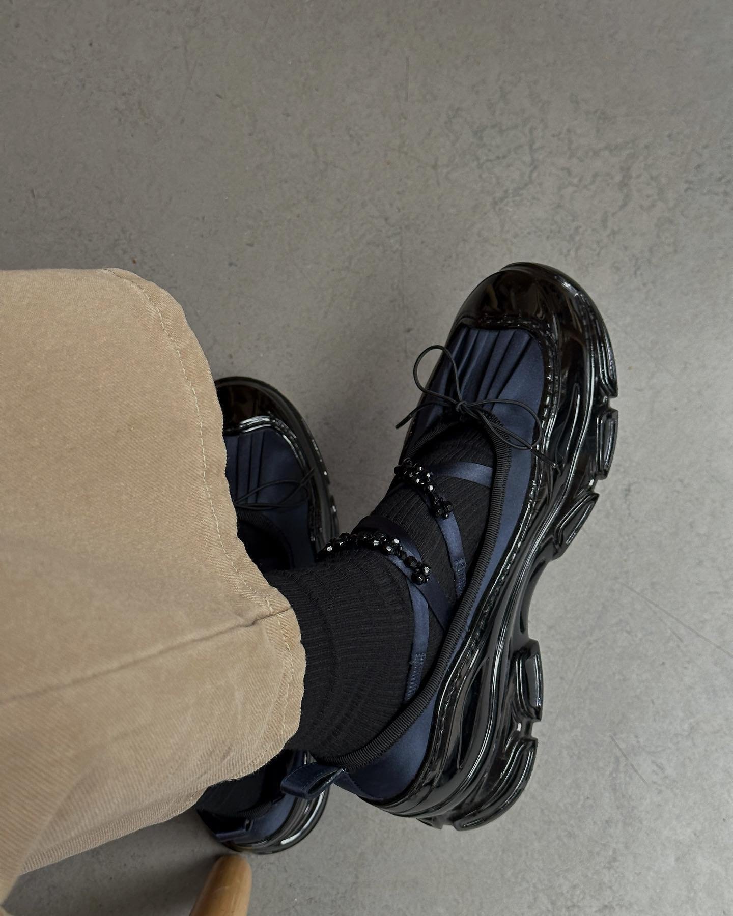 Pessoa com calça bege vestindo sneakers pretos estilizados com solado grosso e meia escura.