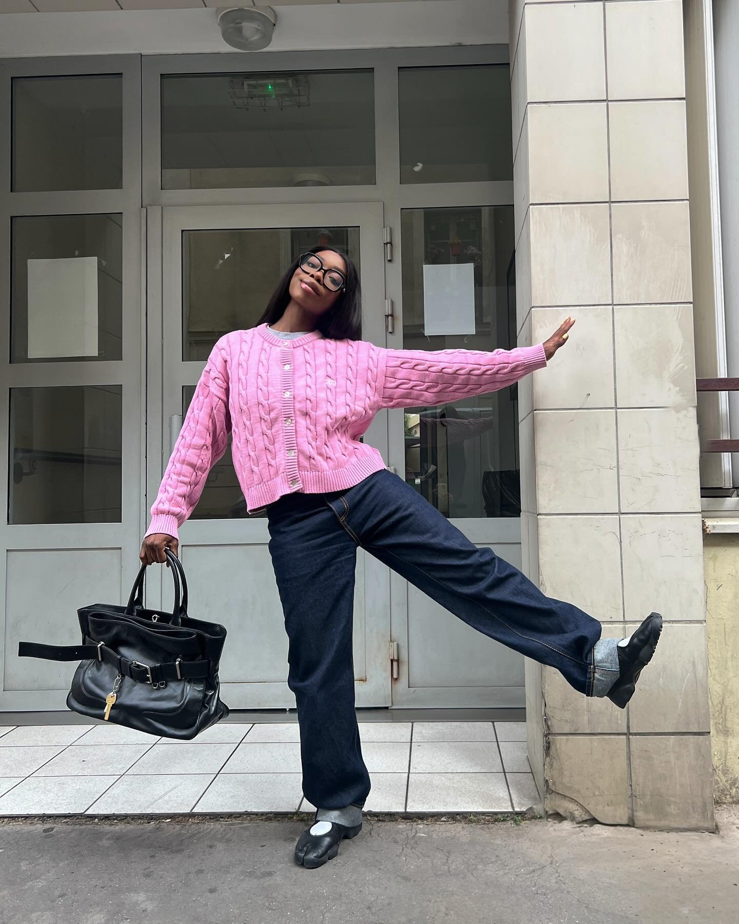 Pessoa sorrindo com suéter rosa e calça jeans, segura bolsa preta. Sneakerina posada, de braços abertos e uma perna erguida.