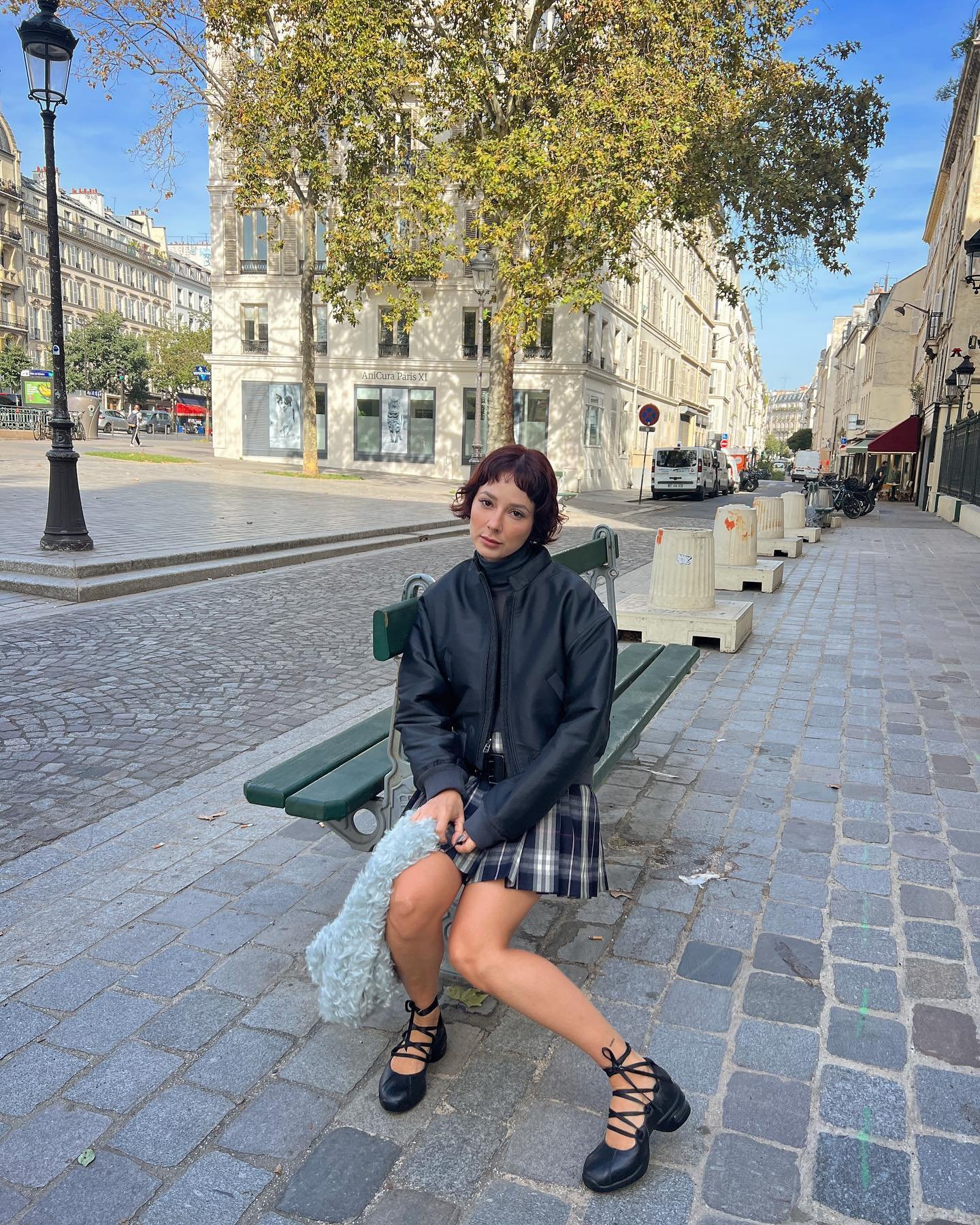 Pessoa sentada em um banco urbano, usando casaco preto e saia xadrez. Rua com prédios e árvores ao fundo.