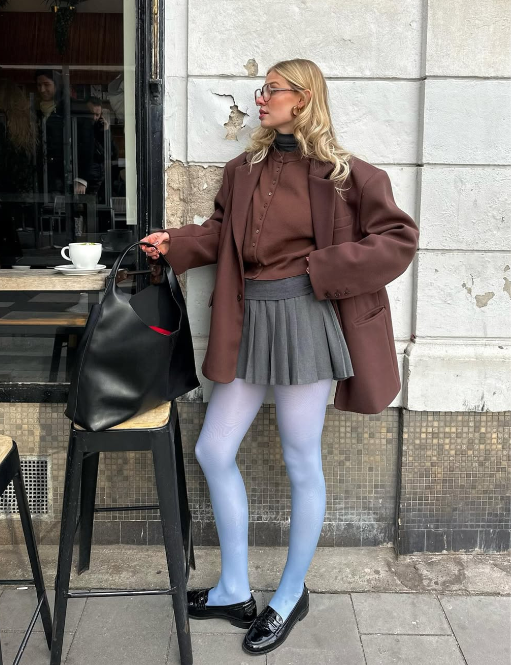 Pessoa com blazer marrom, saia cinza e meia-calça azul, em frente a café, destacando estilo moderno.