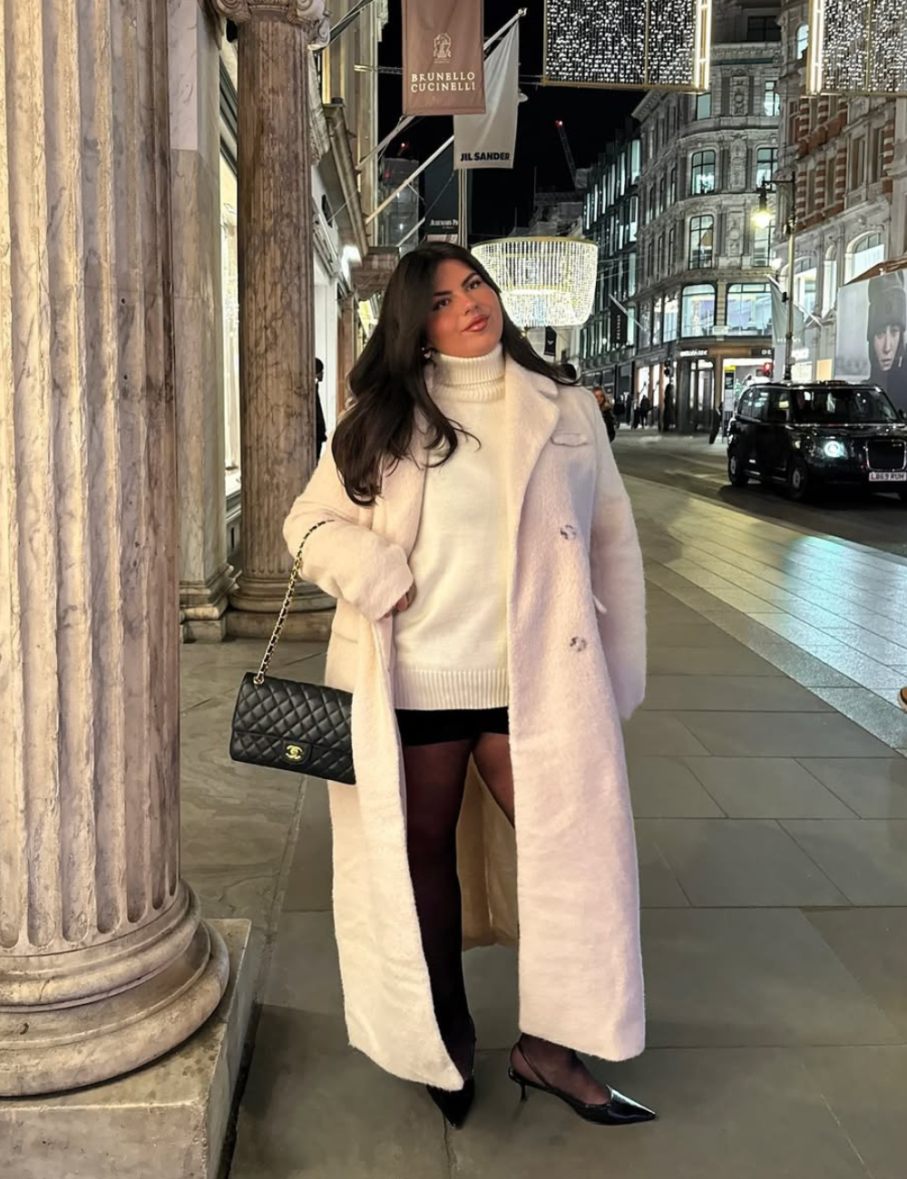 Mulher com casaco rosa e meia-calça preta, em rua iluminada à noite. Segura uma bolsa preta.