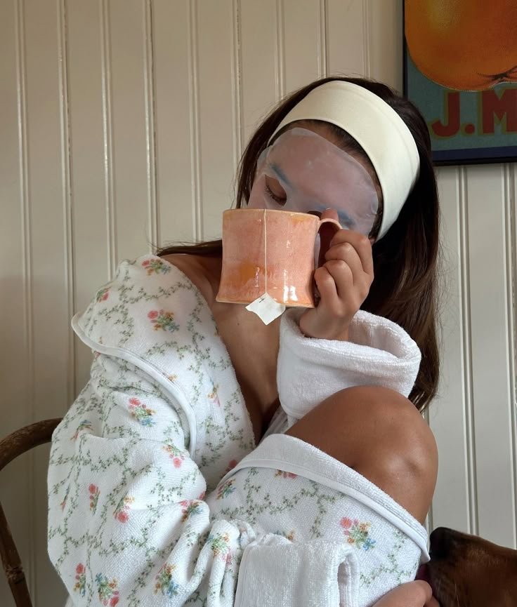 Mulher relaxa com máscara facial e chá em caneca laranja, vestindo roupão floral. Todo dia