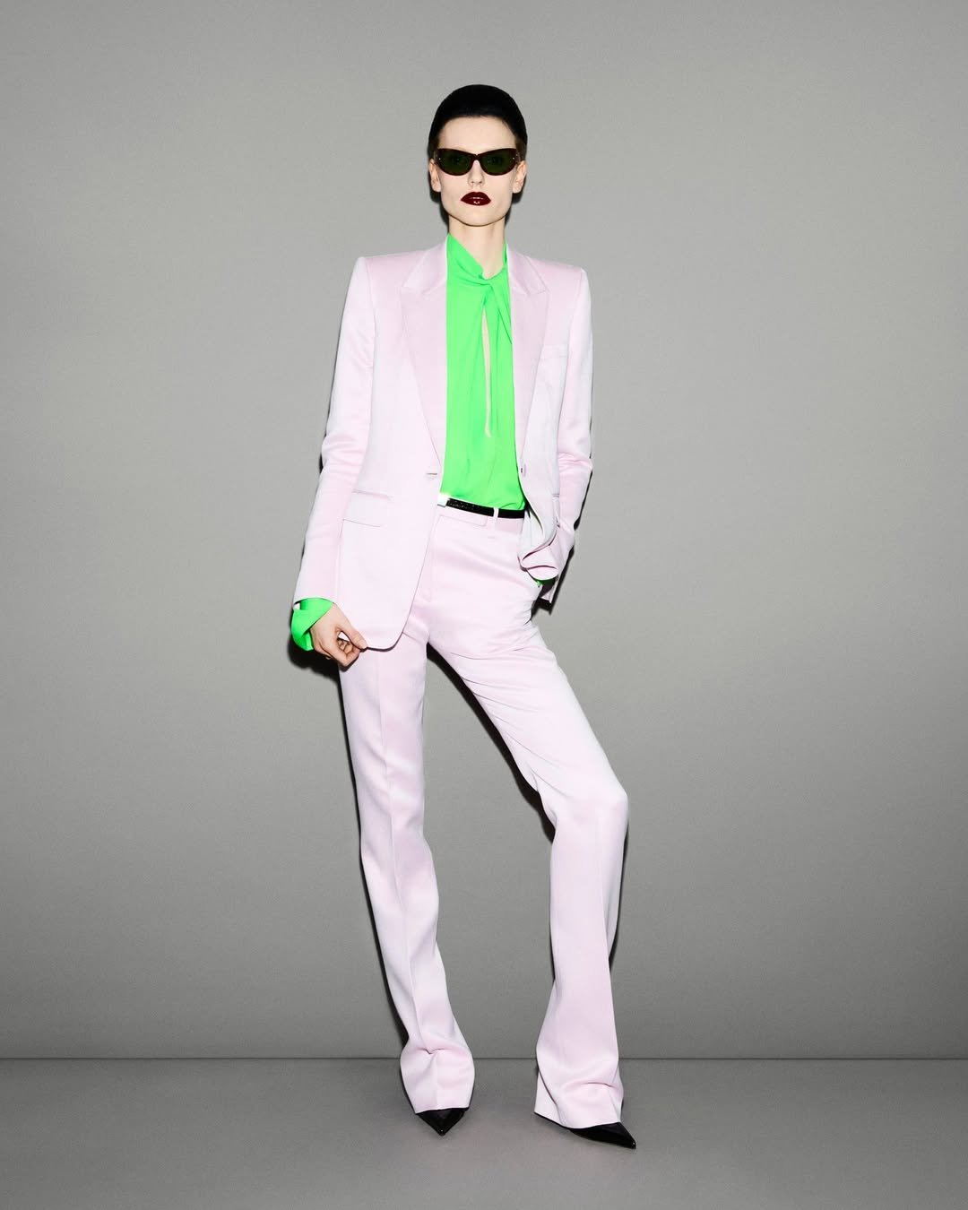 Terno lilás com blusa verde em tons neon, óculos escuros e batom escuro. Fundo cinza neutro.