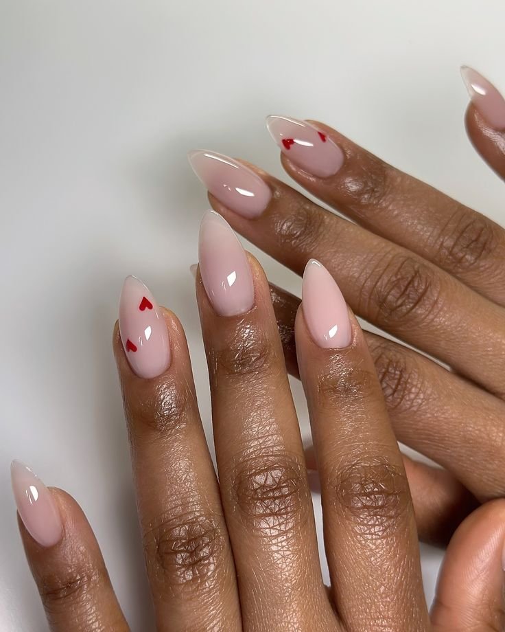Unha amendoada em tom nude com pequenos corações vermelhos no anelar, em mãos de pele escura.