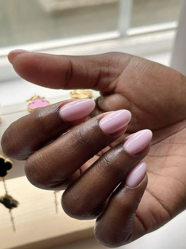 Unha amendoada rosa claro em mão feminina com fundo de joias douradas.
