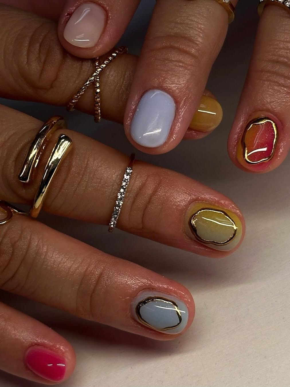 Unhas decoradas em cores variadas com detalhes em dourado, usando anéis finos nos dedos.