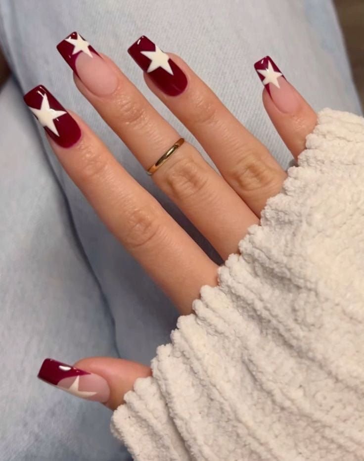 Unhas decoradas em tons de vermelho com estrelas brancas, anel dourado e suéter claro.