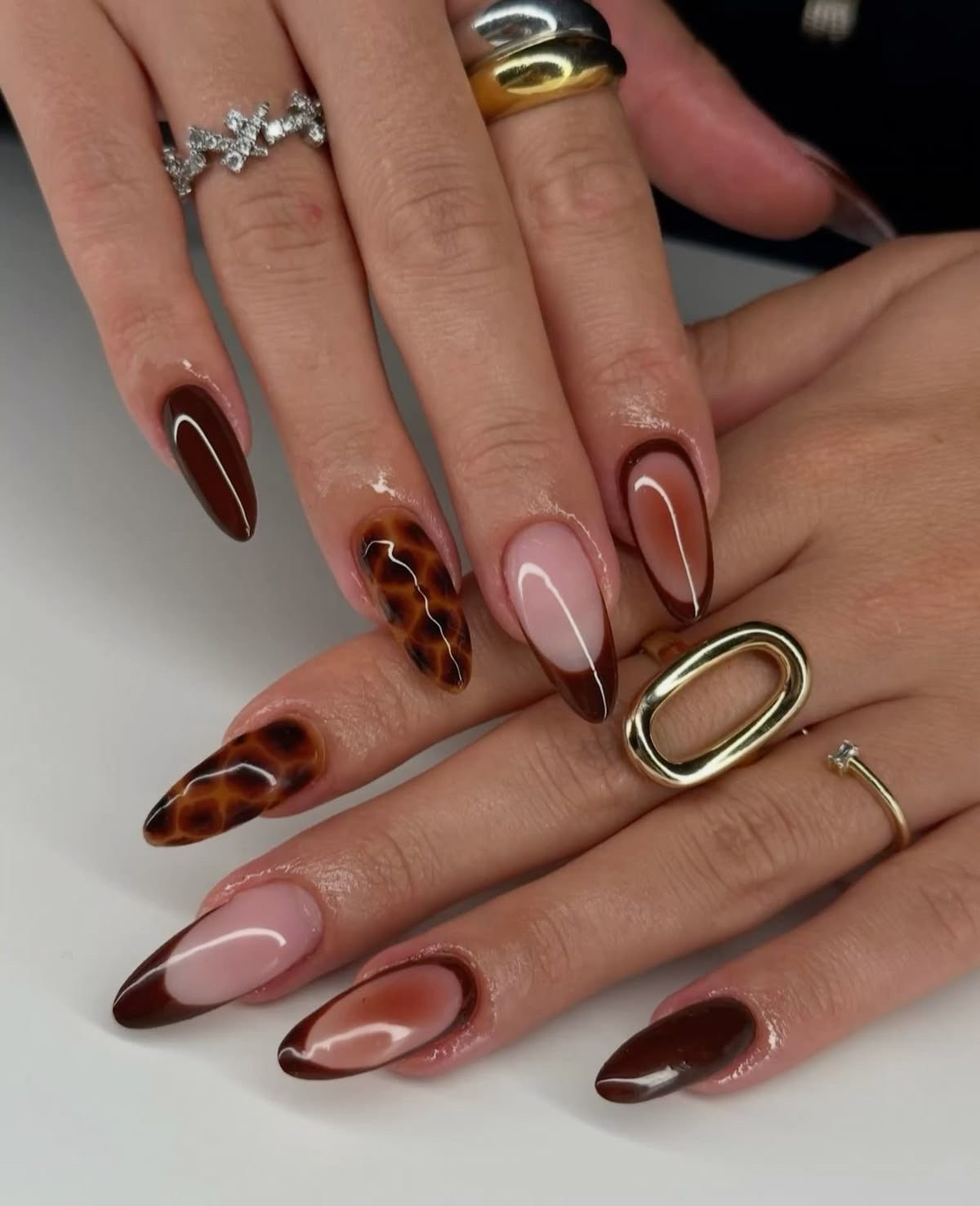 Unhas decoradas com tons de marrom, incluindo efeito tartaruga e degradê, acompanhadas por anéis elegantes.