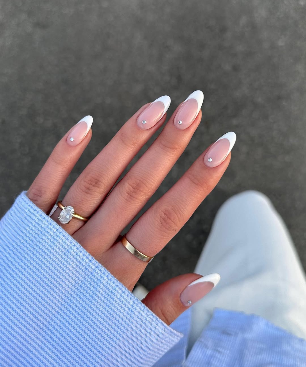 Unhas francesinhas com pedras, estilo elegante para noivas. Mão com anéis sobre camisa azul e fundo desfocado.