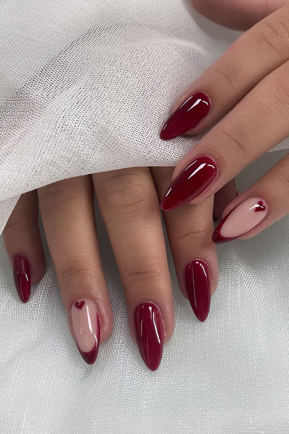 Unhas decoradas em tons de vinho, uma com detalhe de coração discreto.