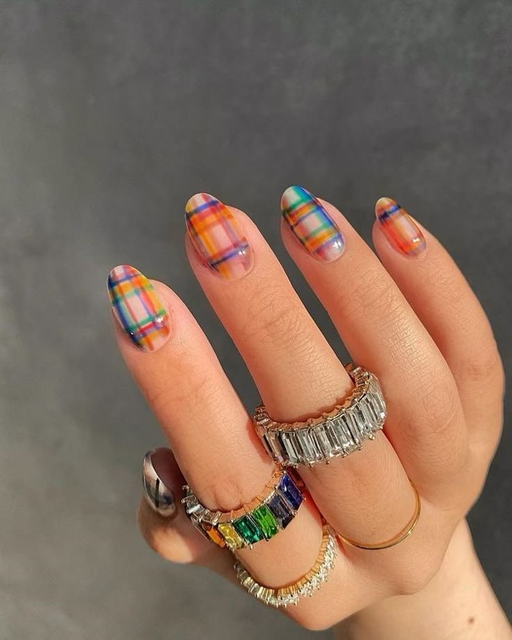 Unhas decoradas com esmalte xadrez colorido em fundo neutro, usando anéis chamativos.