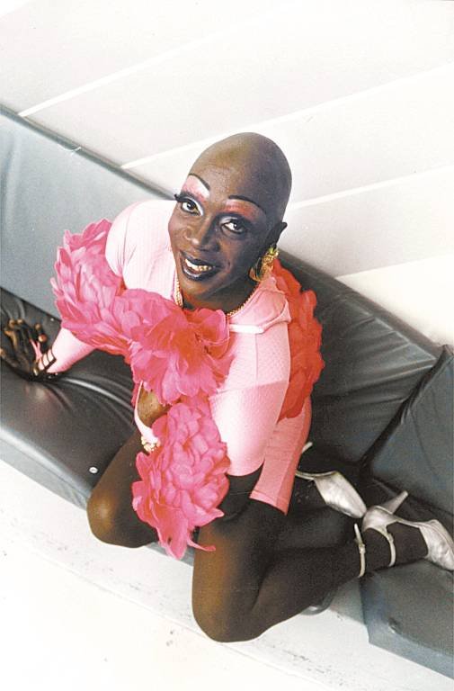 Pessoa sorridente ajoelhada no sofá, vestindo roupa rosa com plumas. Celebrando a diversidade LGBTQIAPN+.