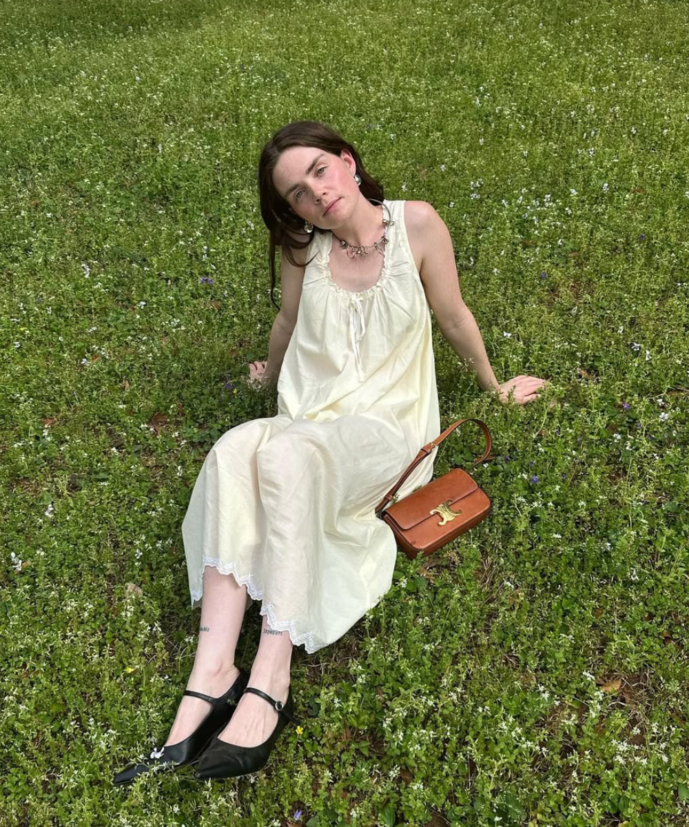 Pessoa sentada na grama usando vestido longo claro, acompanhado de sapatilhas pretas e bolsa marrom ao lado.