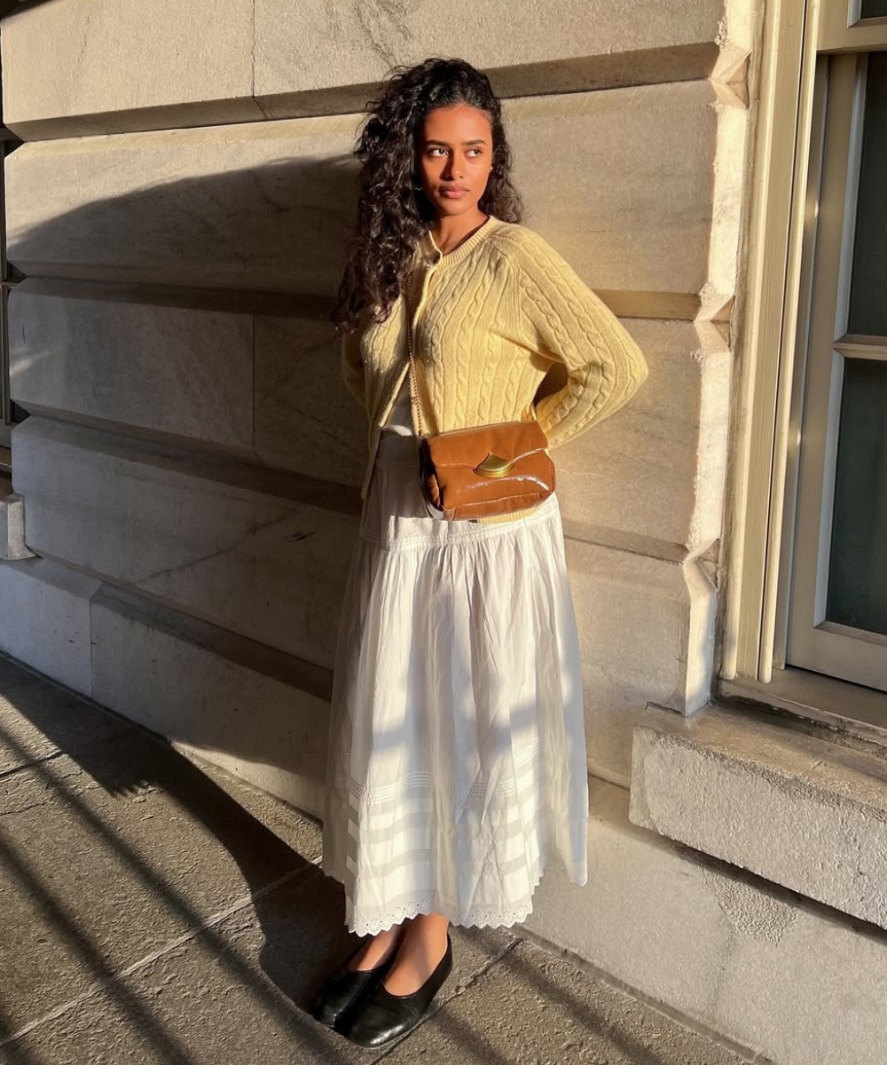 Mulher de suéter amarelo e saia branca, ao sol, segura bolsa marrom. Usa o vestido favorito.