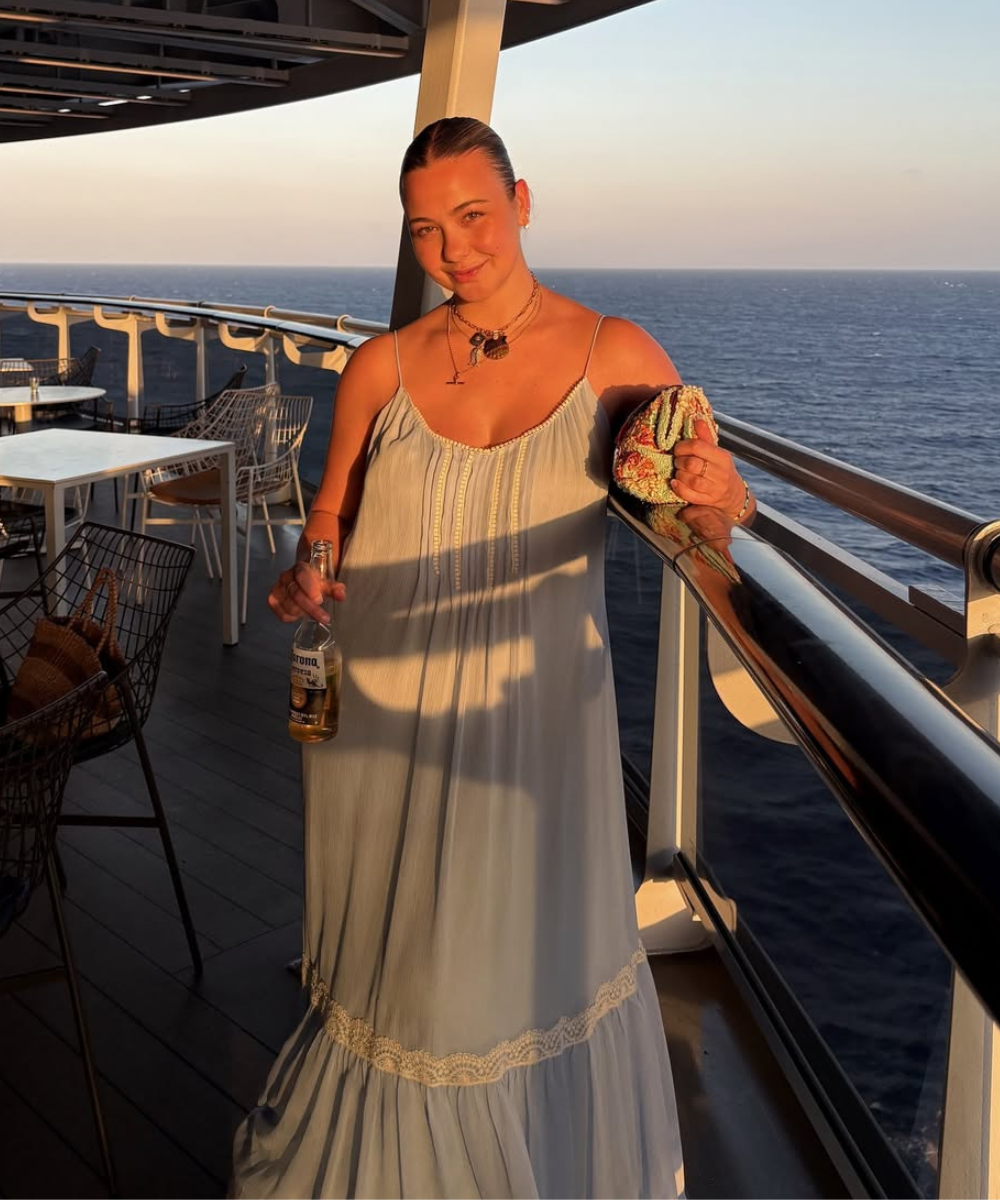 Mulher em varanda de navio ao pôr do sol, usando vestido favorito azul claro e segurando bebida e bolsa.