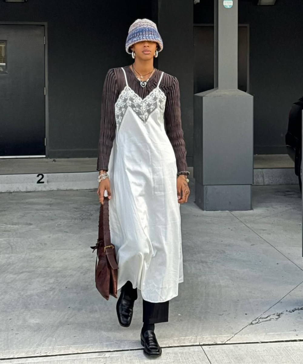 Pessoa usando vestido branco com detalhes rendados sobre blusa preta, chapéu e carregando uma bolsa.